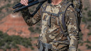 A person wearing Vital S3 Long Sleeve in Valo Camouflage and a large gray backpack, carrying a black tripod over their shoulder in an outdoor desert landscape.