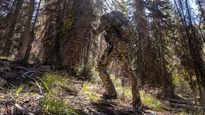 Hunter hiking through tall dry grass on a mountain slope, wearing a Attack Pant in evergreen, with a scenic forested valley in the background.