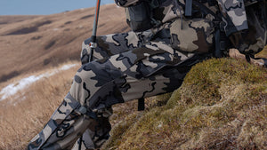 A close-up of a person wearing Attack Pant in Vias camouflage and a matching backpack, sitting on a grassy hillside with a rifle in hand, set against a mountainous outdoor background.