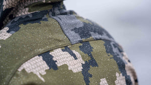 Close-up of a Verde camouflage jacket with a fine textured finish and a blurred light gray background.