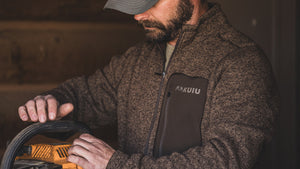 A man wearing a Base Camp Pullover Sweater in Grizzly Heather. He is holding a yellow and black power tool in a dimly lit workshop environment.