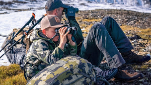 Father and Son in camouflage gear sitting on a rocky landscape both using binoculars.