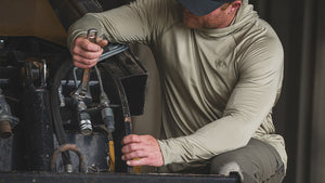 A man wearing Gila Long Sleeve in Talus Grey works on machinery, holding a tool in one hand.