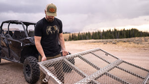 A bearded man wearing a KUIU Camo Big Ram T-Shirt in Valo camouflage stands next to a black UTV in an outdoor desert landscape, holding a large metal mesh cargo bed cover.