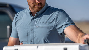 Close‑up of man wearing a Mesa Short Sleeve Shirt in Slate Greyish Blue, standing outdoors beside a vehicle and reaching over a cooler.
