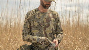 Hunter wearing a ULTRA Merino Valo camouflage long-sleeve shirt and matching camouflage gloves stands in a field of dry golden grass, looking down while holding a small item in his hands.