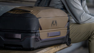 Close-up of a Waypoint 4500 Roller in Bourbon Brown resting on a wooden bench next to a person, Highlighting KUIU ram logo and grab on the side.