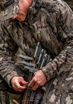 A close-up of a hunter in-full Veros camouflage hunting jacket, pant and cap, packing a bow with string and accessories in a natural outdoor setting.