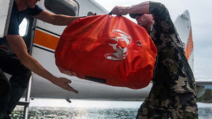 Male hunter in head to toe Verde Camo handing a Blaze Orange KUIU TAKU 9000 Bag to another male in a Super Cub with a lake as the background.
