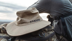 Male hunter in head to toe Verde Camo standing atop lush, green mountains putting his iPhone in his KUIU Phone Holder attached to his Verde Camo PRO Bino Harness.