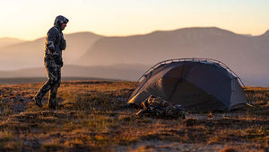 Male hunter in head to toe KUIU Vias Camo walking towards his KUIU Shelter and KUIU PRO Pack with a dusky sunrise in the background and outlines of mountains.