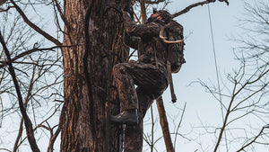 A person in Veros camouflage climbing up a large tree trunk outdoors, with a backpack and wearing hunting gear, with bare branches and a pale sky in the background.