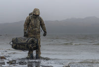 Side of hunter carrying a Taku Blstx Duffel on a rocky beach.