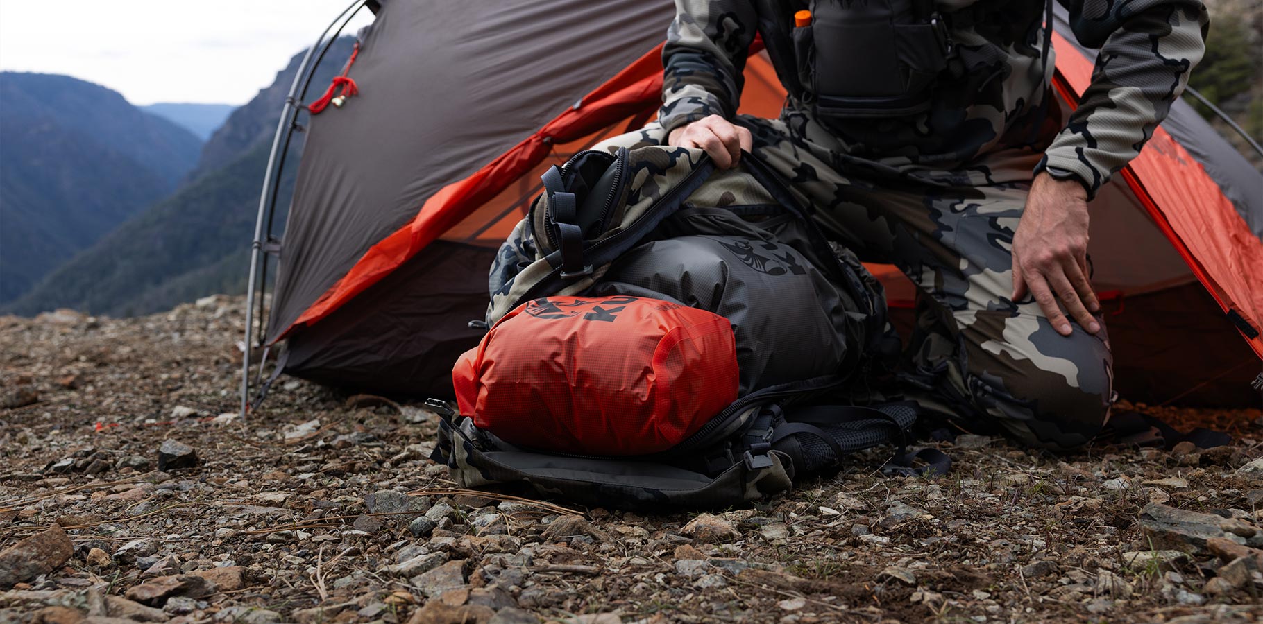 A hunter kneeling outside a tent unpacking his backpack filled with KUIU Roll Top Dry Bags.
