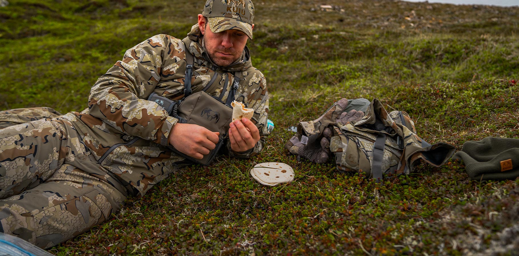 A hunter in Valo Camouflage relaxing on a hillside eating a tortilla.