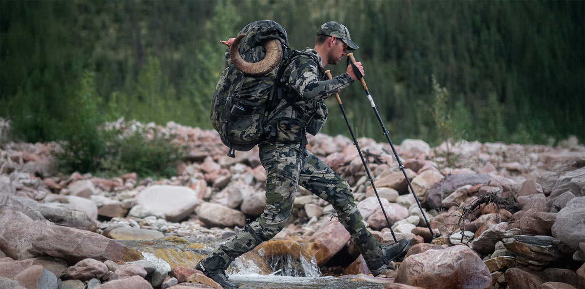 A man hiking through a rocky stream wearing a large Verde camouflage backpack with a ram-horn strapped to the top, camouflage pants, and using trekking poles in an outdoor forest setting.