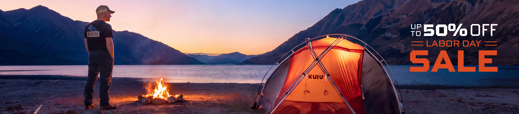 A person stands beside a campfire at dusk next to a bright KUIU orange dome tent pitched on a lakeshore with mountains in the background, and promotional text reading 'UP TO 50% OFF LABOR DAY SALE'.