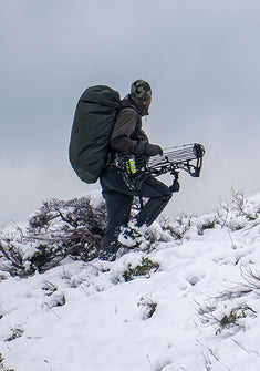Hunter hikes up a snow-covered hill, carrying a large backpack and a compound bow