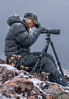 A woman wearing Super Down PRO Hooded Jacket in Gunmetal Grey, sits on a snowy rock, looking through a spotting scope mounted on a tripod in the snow.