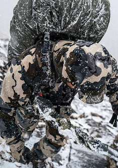 A hunter in Vias camouflage clothes, carrying a large snow-covered backpack while climbing through deep snow.