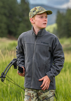 A young boy wearing a Youth Versa Fleece Full Zip Jacket standing in a grassy field and holding a compound bow.