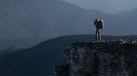 Hunter carrying a large backpack stands on a rocky peak overlooking a mountainous landscape.