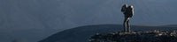 Hunter carrying a large backpack stands on a rocky peak overlooking a mountainous landscape.