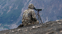 Hunter wearing valo camouflage outerwear looks through a spotting scope while sitting on a rocky ridge with mountains in the background.