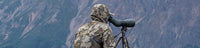 Hunter wearing KUIU valo camouflage outerwear looks through a spotting scope while sitting on a rocky ridge with mountains in the background.