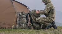 Hunter wearing KUIU valo camouflage outerwear sits beside a dome tent, organizing gear near the tent entrance in an outdoor backcountry setting.