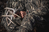 A close-up lifestyle shot of a person in Veros camouflage hunting clothing holding a large antlered hunting trophy.