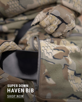 Closeup of a male utilizing his KUIU Super Down Haven's dual entry cargo pockets whether you’re seated or standing. Text reads: Super Down Haven Bib - Shop Now.