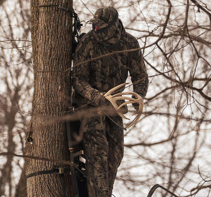 A person in Veros camouflage clothing stands in a tree among bare winter branches, holding a large deer antler or hunting tool.