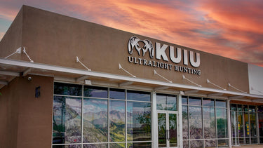 Tempe, Arizona, KUIU Ultralight Hunting storefront exterior at sunset, with a tan building facade, large glass windows, and a dramatic orange-pink sky reflected in the windows.