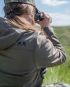 Hunter Using binoculars sitting down looking away from the camera while wearing the Women's Guide DCS Jacket in Ash Brown