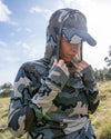 Female hunter wearing Women's Gila LS Hoodie in Vias Camouflage with hands on hood near neck